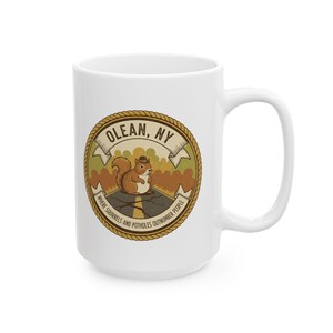 May include: White ceramic mug featuring a circular design with the text "OLEAN, NY" and a cartoon squirrel wearing a hat. The design also includes the phrase "WHERE SQUIRRELS AND POTHOLES OUTNUMBER PEOPLE."