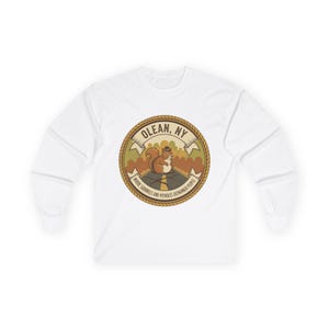 May include: White long-sleeve shirt featuring a circular graphic with a squirrel wearing a hat, the text "OLEAN, NY", and the phrase "WHERE SQUIRRELS AND POTHOLES OUTNUMBER PEOPLE". The design is in shades of brown, tan, and green.