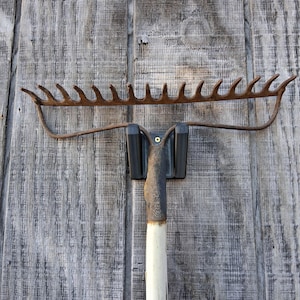 May include: A vintage garden rake with a rusty, curved metal head and tines. The handle is white with a black grip. The rake is set against a weathered, gray wooden background.