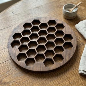 May include: A round, dark brown wooden trivet with a honeycomb pattern. The trivet is on a wooden table, with a small ceramic bowl and a wooden spoon to the side. A linen napkin is also visible.