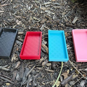 May include: Four rectangular trays in black, red, turquoise, and pink. The trays are made of plastic and have textured surfaces. The trays are arranged in a row on a bed of wood chips.