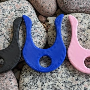 May include: Three U-shaped, 3D-printed objects in black, blue, and pink, each with a central hole. The objects are arranged on a bed of gray and pink stones, showcasing a minimalist design.