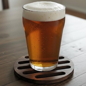 May include: A pint glass filled with amber-colored beer and a thick white head, resting on a dark wooden coaster with a slotted design. The glass has condensation droplets on the outside.