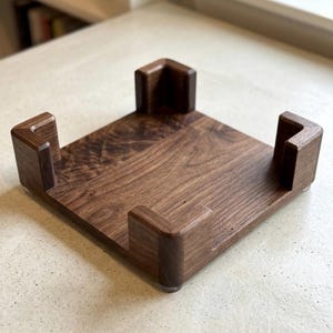 May include: A square, wooden stand with a rich, dark brown finish. The stand features four corner supports and small, clear feet. The wood grain is visible, adding to the natural aesthetic. This item is likely designed to hold a record player or other electronic device.