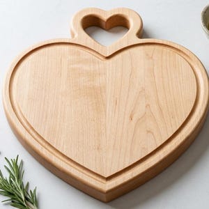 May include: A heart-shaped wooden cutting board with a handle featuring a heart-shaped cutout. The light-colored wood has a groove around the edge. A small bowl of olive oil and herbs, peppercorns, and rosemary sprigs are also visible.