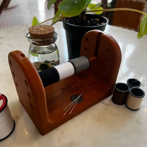 May include: A wooden thread holder with a JMC logo, four spools of thread, and four needles. The holder has a slot for holding spools of thread and a small compartment for needles.