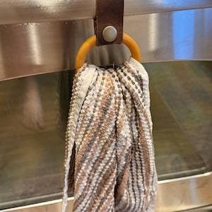 May include: A kitchen towel hanging from a brown leather strap with a wooden ring. The towel has a striped pattern in shades of brown, beige, and white. The strap is attached to a metal oven handle with a silver button.