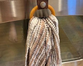 May include: A kitchen towel hanging from a brown leather strap with a wooden ring. The towel has a striped pattern in shades of brown, beige, and white. The strap is attached to a metal oven handle with a silver button.