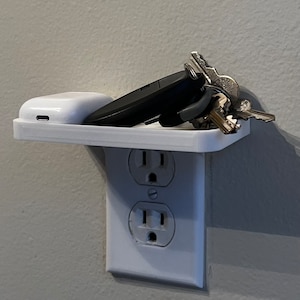 Puede incluir: Una estantería de plástico blanco montada sobre una toma de corriente estándar. La estantería contiene un juego de llaves, una funda negra para auriculares inalámbricos y una funda de carga blanca.