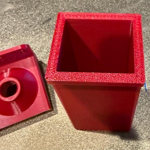 May include: A red, square-shaped container with a textured rim and a separate red piece with a circular hole. The container has a tapered design, and the detached piece appears to be a lid or base. The items are made of a solid, opaque material.