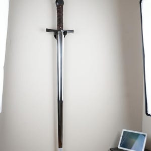 May include: A long, double-edged steel sword with a dark brown leather-wrapped grip, a rounded pommel, and a straight crossguard. The sword is mounted vertically on a wall using a black display bracket, featuring a polished blade and a silver tip.