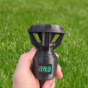 May include: A black, 3D-printed device with a digital display showing the number 8.8.8 in green. The device has a unique, geometric design with a flared top and is held against a backdrop of green grass.
