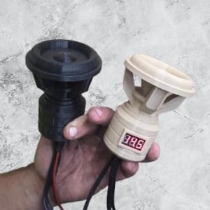 May include: Two small, 3D-printed speaker enclosures. One is black, the other is beige. The beige speaker has a digital display showing "385". Both have wires extending from the base. Held in a hand against a mottled gray background.