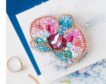 Può includere: Una spilla artigianale con un vivace design floreale composto da perline rosa, blu e turchesi. Il pezzo è accentuato da cristalli sfaccettati rosa e trasparenti, con perline color oro che delineano i bordi delle forme dei petali.