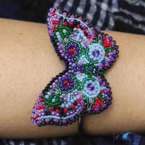 May include: A beaded butterfly bracelet with purple, green, red, and white beads. The bracelet is on a person's wrist.