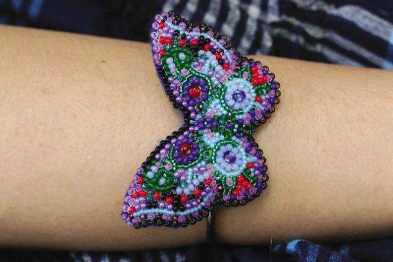 May include: A beaded butterfly bracelet with purple, green, red, and white beads. The bracelet is on a person's wrist.