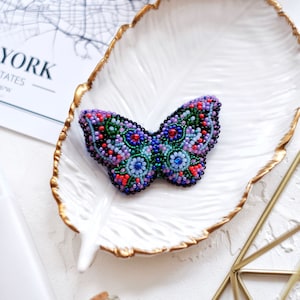 May include: A beaded butterfly brooch with a purple, blue, green, and red color scheme. The brooch is sitting on a white plate with a gold rim.