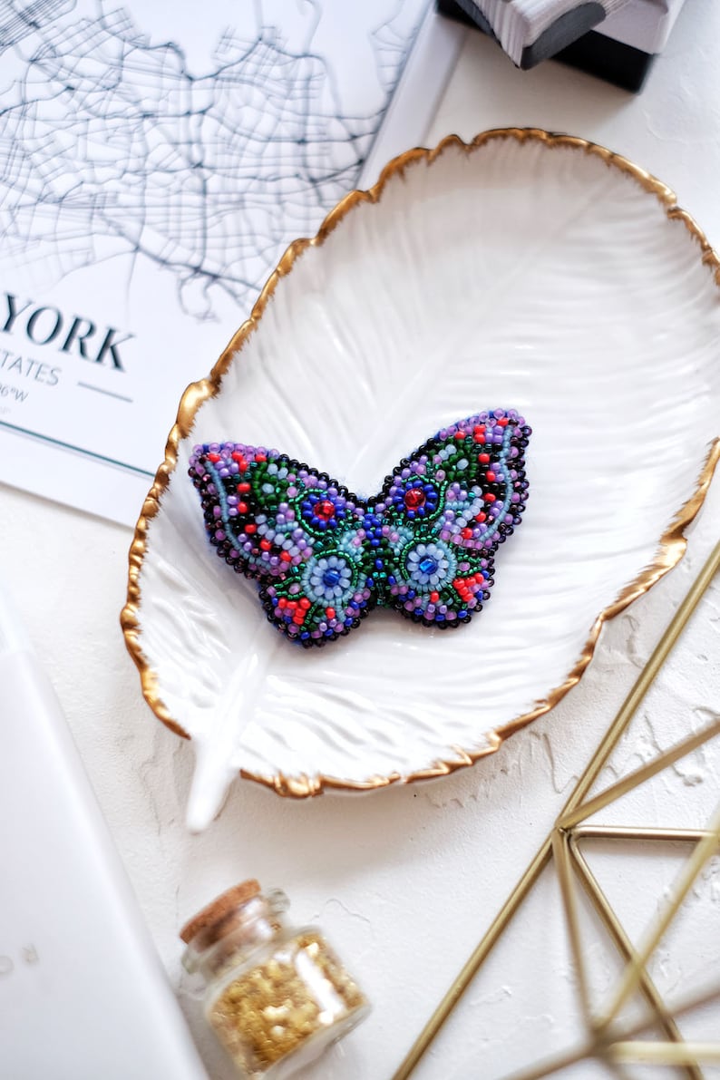 May include: A beaded butterfly brooch with a purple, blue, green, and red color scheme. The brooch is sitting on a white plate with a gold rim.