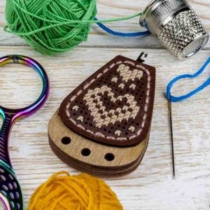 May include: A wooden needle minder with a brown leather top featuring a cross-stitch design of hearts. The minder has three holes for needles and is surrounded by green, blue, and orange yarn.