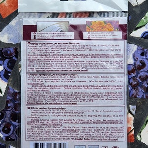 May include: A close-up of the back of a packaging for a bead embroidery kit. The packaging is white with blue and red accents. The text on the packaging reads "Kit-decoration for embroidery" and includes a list of the kit's contents, such as beads, needles, felt, and instructions. The packaging also includes a warning that the kit is not suitable for children under 3 years old.