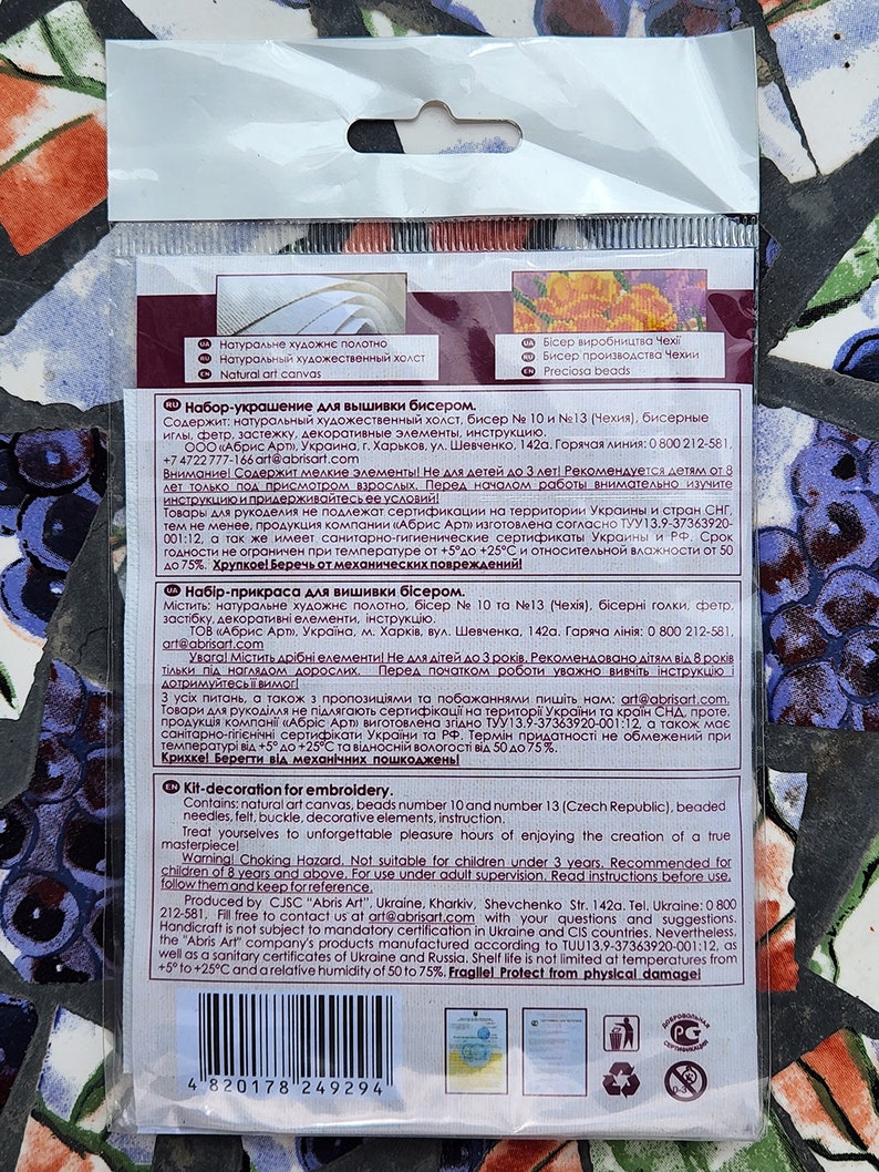 May include: A close-up of the back of a packaging for a bead embroidery kit. The packaging is white with blue and red accents. The text on the packaging reads "Kit-decoration for embroidery" and includes a list of the kit's contents, such as beads, needles, felt, and instructions. The packaging also includes a warning that the kit is not suitable for children under 3 years old.