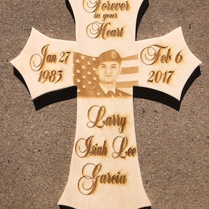 May include: Wooden cross-shaped memorial with a photo of a soldier and an American flag. The cross features the dates Jan 27, 1983 and Feb 6, 2017, along with the name Larry Isiah Lee Garcia and the phrase "Forever in your Heart."
