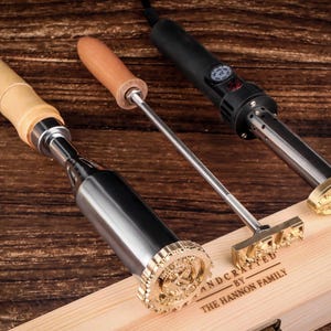 以下が含まれることがあります： 真鍮スタンプと木製ハンドルが付いた3つの木材焼印が、木製の表面に表示されています。「HANDCRAFTED BY THE HANNON FAMILY」の文字が木箱に表示されています。焼印には黒いコードが付いています。