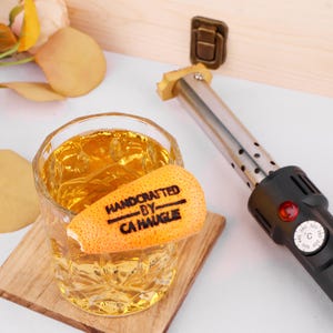Könnte beinhalten: Nahaufnahme eines Glases mit bernsteinfarbener Flüssigkeit und einer kleinen Orangenschalen-Garnitur. Die Schale trägt die eingebrannten Worte "HANDCRAFTED BY CA HAUGLE". Im Hintergrund ist ein schwarz-silbernes elektrisches Branding-Eisen zu sehen.