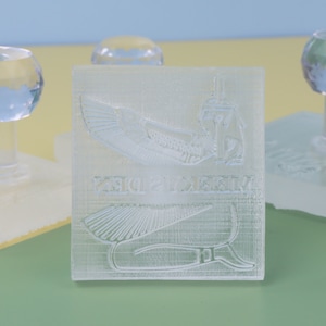 Puede incluir: Un sello de pl&aacute;stico transparente con un dise&ntilde;o en relieve de una diosa egipcia con alas y jerogl&iacute;ficos. El sello est&aacute; sobre una base blanca.