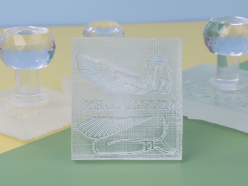 Puede incluir: Un sello de pl&aacute;stico transparente con un dise&ntilde;o en relieve de una diosa egipcia con alas y jerogl&iacute;ficos. El sello est&aacute; sobre una base blanca.
