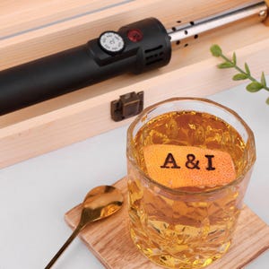 Pode incluir: Um close-up de um copo de líquido âmbar com uma casca de laranja flutuando no topo. A casca tem as letras "A & I" queimadas.