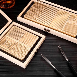 Puede incluir: Dos cajas de madera con insertos de metal de color dorado. Los insertos tienen diferentes patrones geométricos. Las cajas están abiertas y los insertos son visibles. Hay dos herramientas de metal sobre una superficie negra.