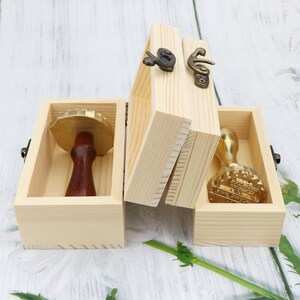Puede incluir: Dos cajas de madera con bisagras y cierres de lat&oacute;n. Cada caja contiene un sello de lat&oacute;n con un mango de madera. Un sello tiene un dise&ntilde;o decorativo y el otro tiene una superficie en blanco.