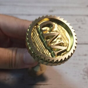 Puede incluir: Un sello de metal dorado con un borde dentado y las letras "G.W." en relieve en el centro.