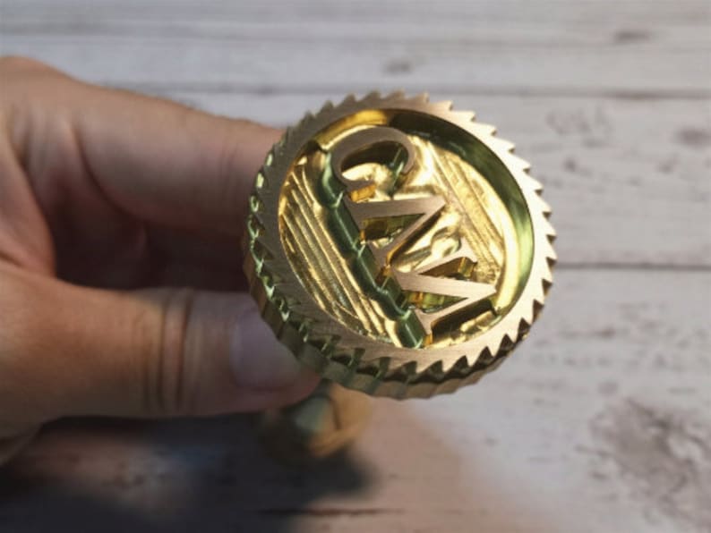 Puede incluir: Un sello de metal dorado con un borde dentado y las letras "G.W." en relieve en el centro.