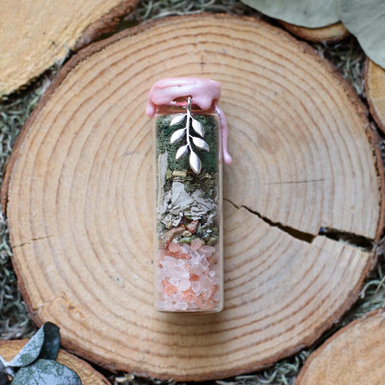 May include: A small glass bottle filled with layers of pink crystals, dried herbs, and green material. The bottle is sealed with pink wax and a silver leaf charm. The bottle is set on a wooden slice.