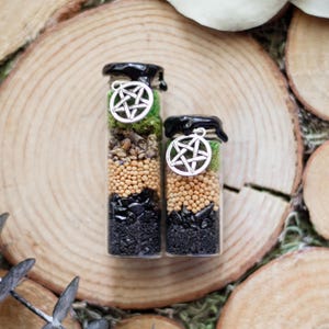 May include: Two small glass bottles with black lids, filled with layers of ingredients. The layers include black stones, small tan beads, and green moss. Each bottle has a silver pentagram charm hanging from the lid. The bottles are on a wooden surface.