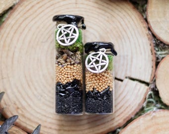 May include: Two small glass bottles with black lids, filled with layers of ingredients. The layers include black stones, small tan beads, and green moss. Each bottle has a silver pentagram charm hanging from the lid. The bottles are on a wooden surface.