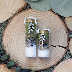 May include: Two glass bottles filled with layers of white, green, and purple materials. The bottles have gold-colored lids with white wax drippings and a silver leaf charm. The bottles are on a wooden slice.