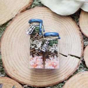 May include: Two clear glass vials filled with layers of pink salt, brown stones, dried herbs, and green moss. Each vial is sealed with blue wax and features a silver-toned lantern charm tied around the neck with thin twine.