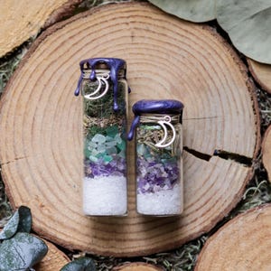 May include: Two glass bottles filled with layers of white, green, and purple crystals and herbs. The bottles are sealed with purple wax and adorned with a silver crescent moon charm. The bottles are displayed on a wooden surface.