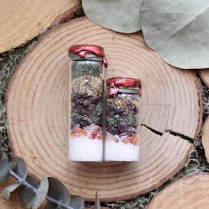 May include: Two glass jars filled with layers of ingredients, including herbs, crystals, and white granules. The jars are sealed with red wax and sit on a wooden surface, suggesting a natural or mystical theme.