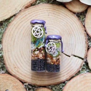 May include: Two glass bottles filled with layers of black, brown, and green materials, topped with purple wax and a silver pentacle charm. The bottles are displayed on a wooden surface, creating a natural aesthetic.