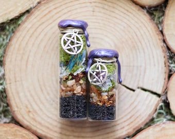 Könnte beinhalten: Zwei Glasflaschen gefüllt mit Schichten aus schwarzen, braunen und grünen Materialien, gekrönt mit violettem Wachs und einem silbernen Pentagramm-Anhänger. Die Flaschen werden auf einer Holzoberfläche präsentiert, was eine natürliche Ästhetik schafft.