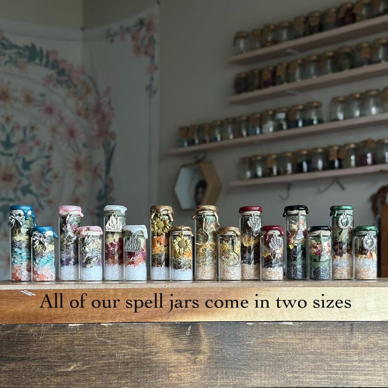 May include: A collection of small glass spell jars filled with colorful crystals, dried flowers, and herbs, each sealed with wax and adorned with a metal charm. The jars are arranged in a row on a wooden surface with the text All of our spell jars come in two sizes.