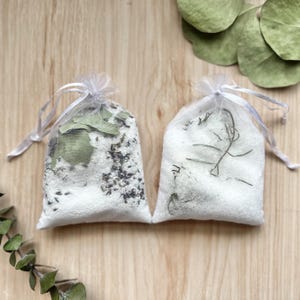 Puede incluir: Dos bolsas de malla blanca llenas de sales de baño y hierbas secas. La bolsa de la izquierda contiene lavanda y eucalipto, mientras que la bolsa de la derecha contiene romero.
