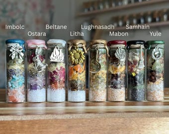 May include: Eight clear glass vials filled with layered botanical materials, crystals, and salts, each sealed with colored wax. Charms include a bow, rabbit, fire, sunflower, crescent moon, wheat sheaf, broom, and wreath, labeled Imbolc, Ostara, Beltane, Litha, Lughnasadh, Mabon, Samhain, and Yule.