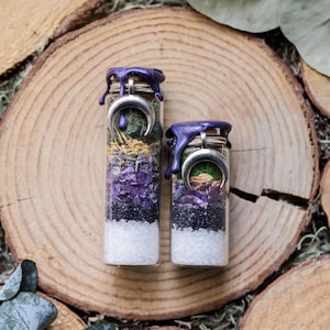 May include: Two glass jars filled with layers of white, black, purple, and green materials. Each jar is topped with a silver crescent moon charm and sealed with purple wax. The jars are set on a wooden slice.