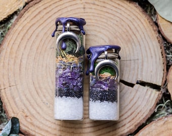May include: Two glass jars filled with layers of white, black, purple, and green materials. Each jar is topped with a silver crescent moon charm and sealed with purple wax. The jars are set on a wooden slice.