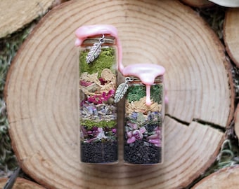 May include: Two small glass bottles filled with layers of colourful dried herbs, flowers, and crystals. Each bottle is topped with a pink wax seal and a silver feather charm. The bottles are displayed on a wooden surface.